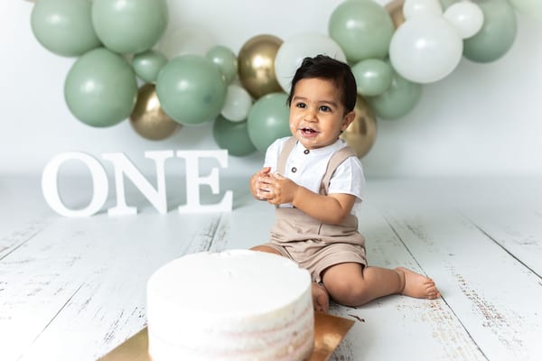 Lachender Junge beim Cake Smash vor salbeigrünen und goldenen Ballons, daneben die Torte
