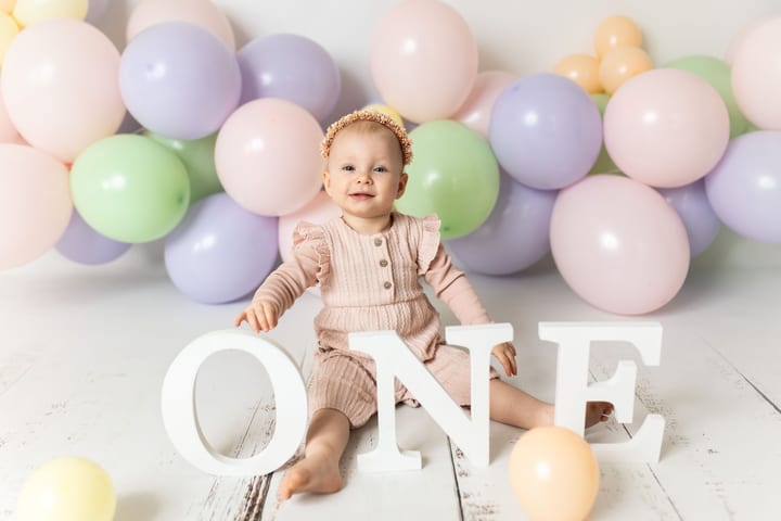 Einjähriges Mädchen sitzt zwischen Pastellballons hinter den Buchstaben ONE