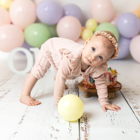 Geburtstagskind krabbelt vor Pastellballons auf den Ball zu, daneben die Torte