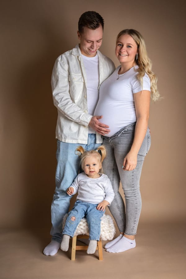 Eltern stehen vor braunem Hintergrund, die kleine Tochter sitzt vor ihnen auf einem Hocker