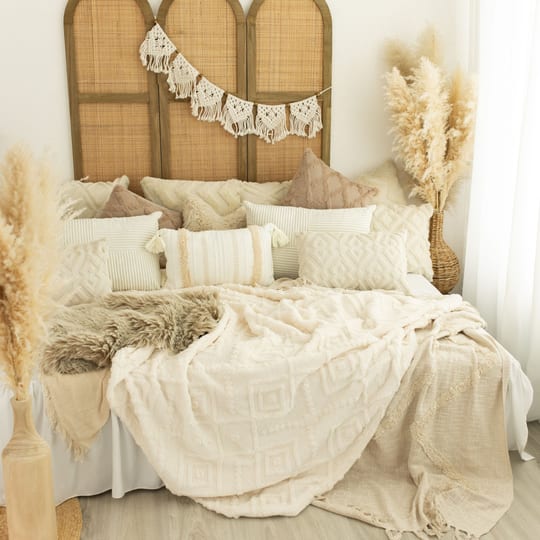 Atelier-Ecke mit Bett, hellen Kissen, Decken und Pampasgras vor einem Rattan-Paravent