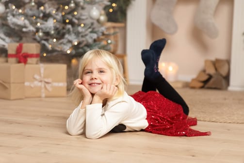 Mädchen im roten Paillettenrock liegt auf dem Bauch vor Tannenbaum und Geschenken