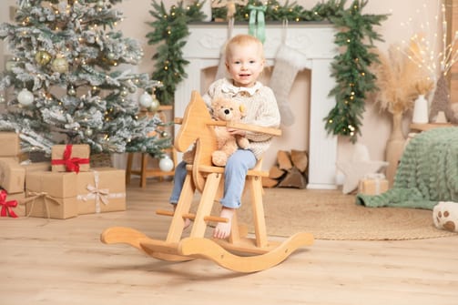 Kleinkind mit Teddy sitzt auf einem Holz-Schaukelpferd vor dem Weihnachtsbaum