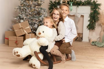 Drei lachende Geschwister auf einem Holzschlitten mit großem Teddy vor dem Kamin