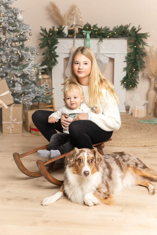 Große Schwester hält ihr kleines Geschwisterchen auf einem Schlitten, davor liegt ein Australian Shepherd