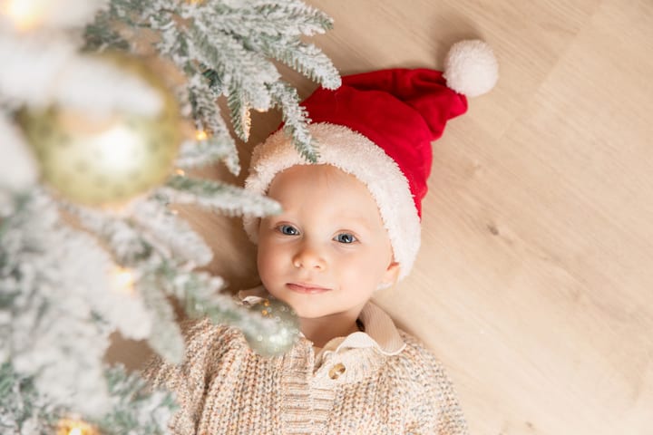 Baby mit roter Weihnachtsmütze liegt neben einem verschneiten Tannenzweig auf dem Holzboden